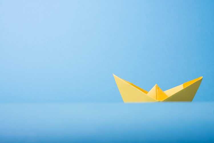 Yellow Paper Folded into Shape of Boat Against Light Blue Paper Background